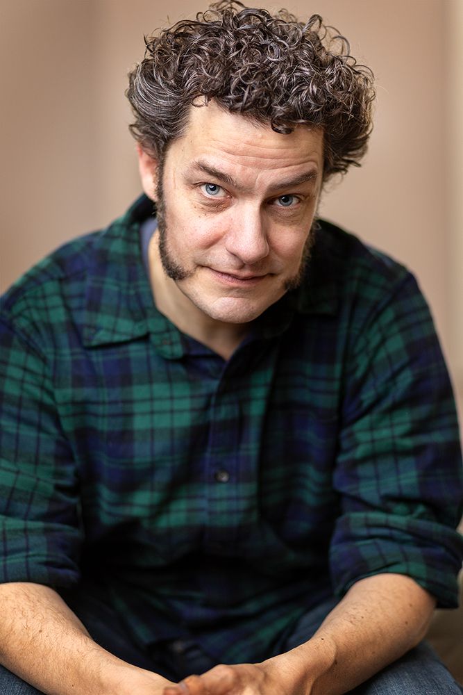A white man with brown curly hair and mutton chop sideburns sits in a checked shirt and jeans with his hands casually held together in front of him