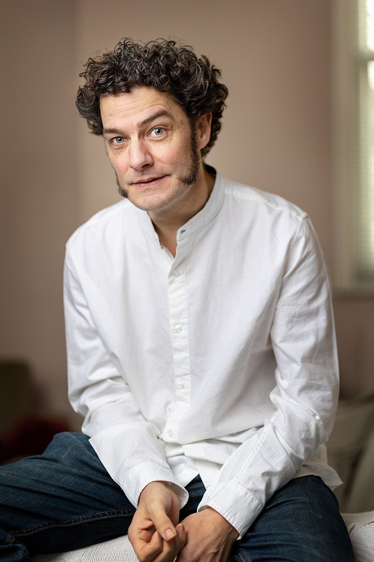 A white man with brown curly hair and mutton chop sideburns sits in a white shirt and jeans with his hands casually held together in front of him