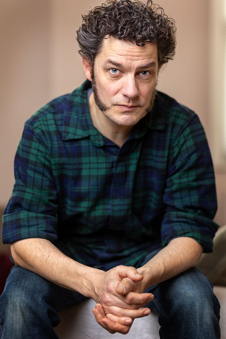 A white man with brown curly hair and mutton chop sideburns sits in a checked shirt and jeans with his hands casually held together in front of him